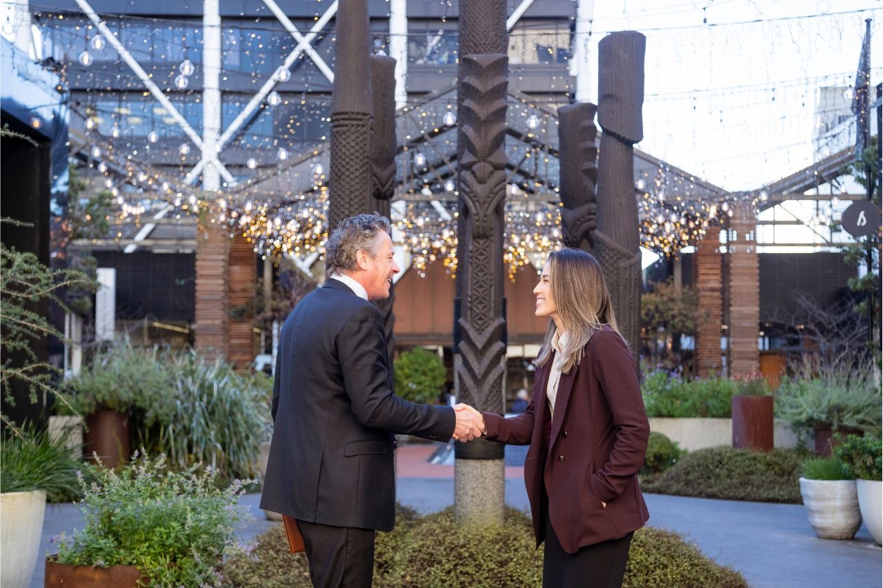 Two business professionals shaking hands in an outdoor Auckland venue featuring Māori carvings, symbolising partnerships supported by the Auckland Business Events Fund.