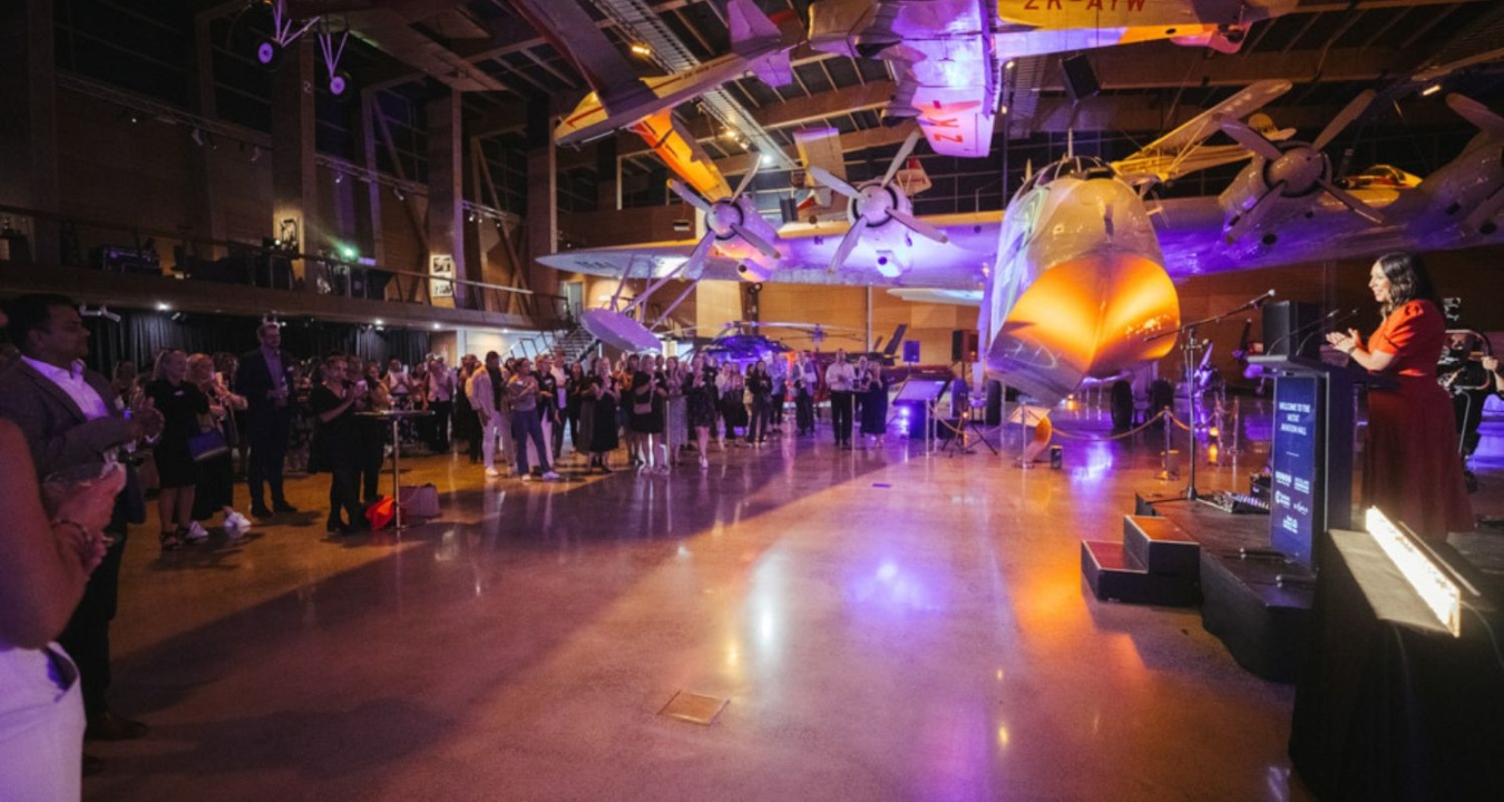 A group listen to a speaker in an orange dress at an event in the MOTAT aviation hall