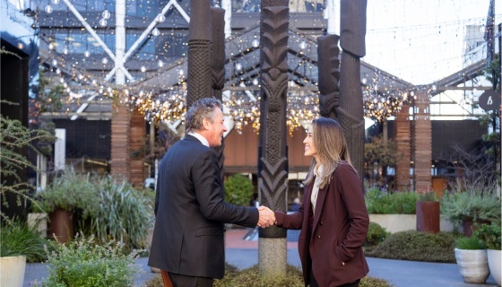 Two business professionals shaking hands in an outdoor Auckland venue featuring Māori carvings, symbolising partnerships supported by the Auckland Business Events Fund.