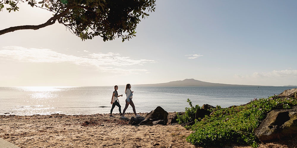 Two women walking along a beach in Auckland with Rangitoto Island in the background